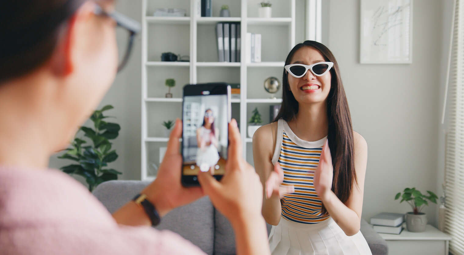 Jonge vrouw met zonnebril danst en lacht terwijl een ander haar filmt met een smartphone in een woonkamer.